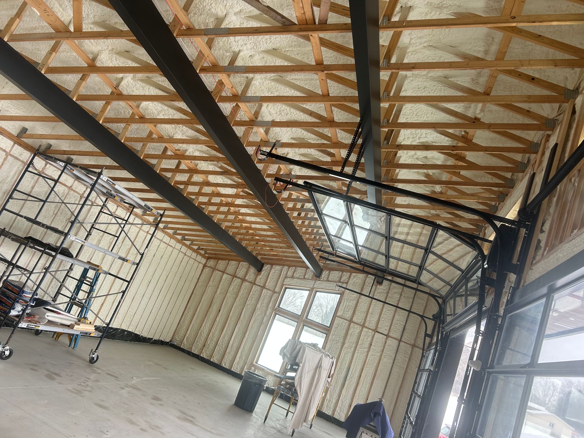 Interior of a building under construction, showing insulated walls and ceiling, glass garage door, and scaffolding.