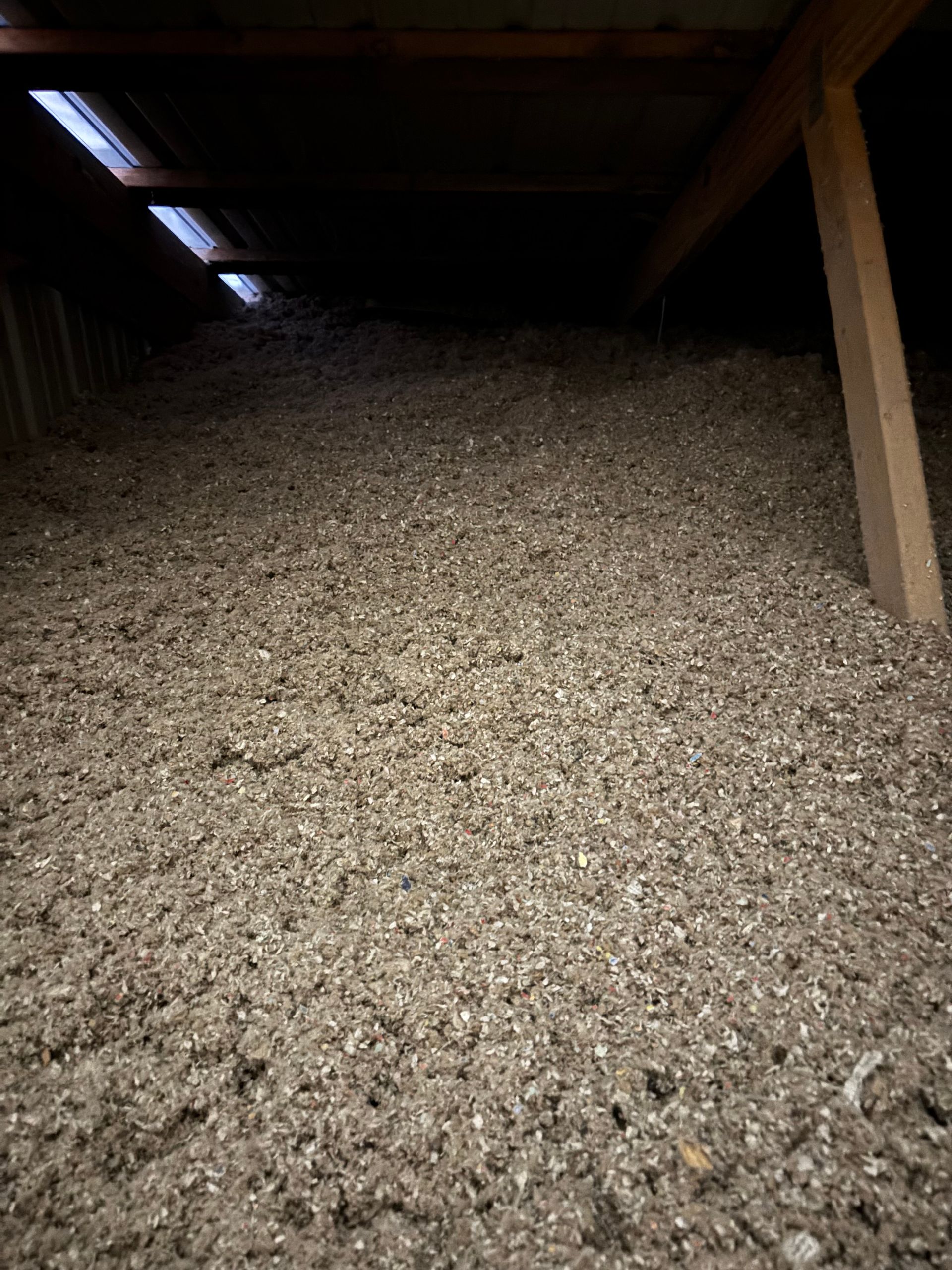 Attic filled with loose fill insulation. Beige and gray material covers the floor under wooden beams and a small skylight.