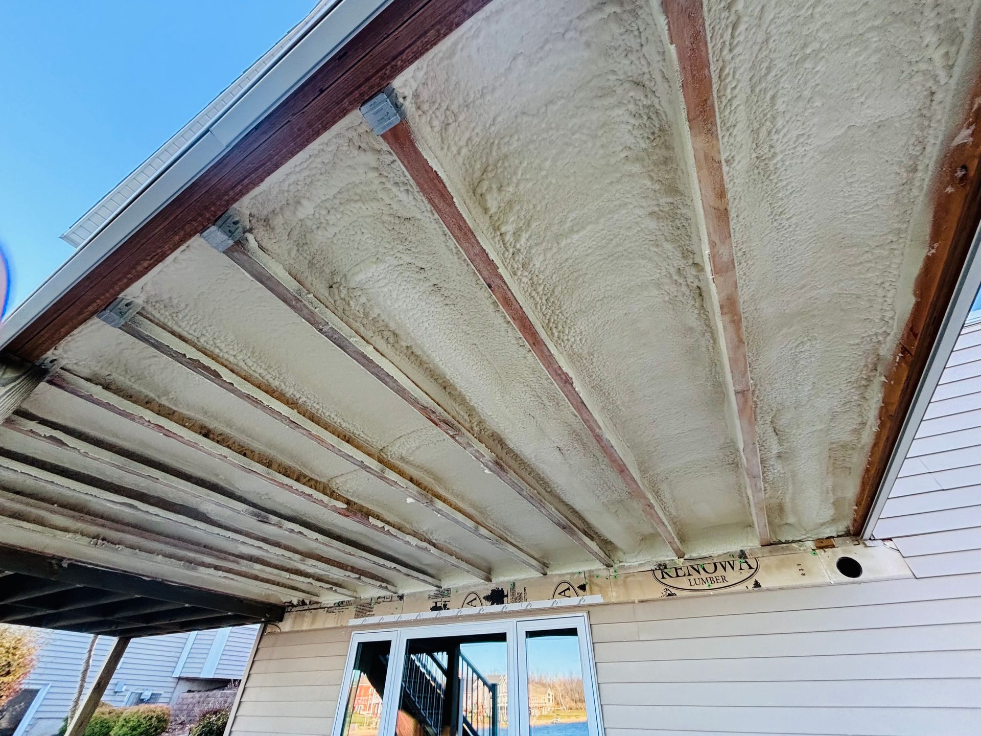 View of a ceiling with spray foam insulation between wooden beams, viewed from below a porch.