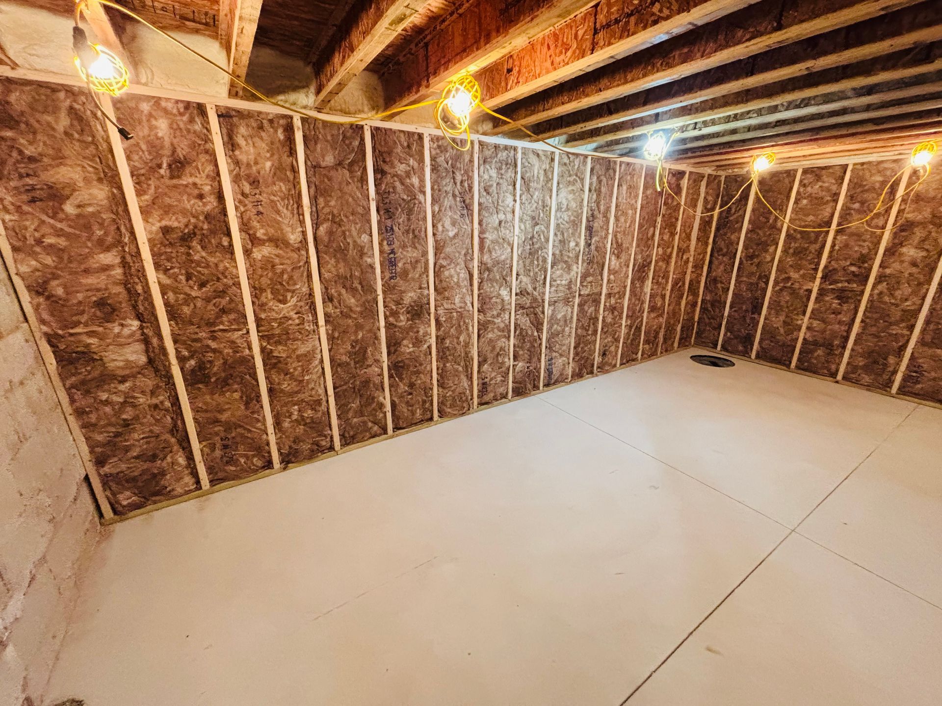 Basement interior construction, insulation installed on wooden studs. Concrete floor and exposed ceiling beams.