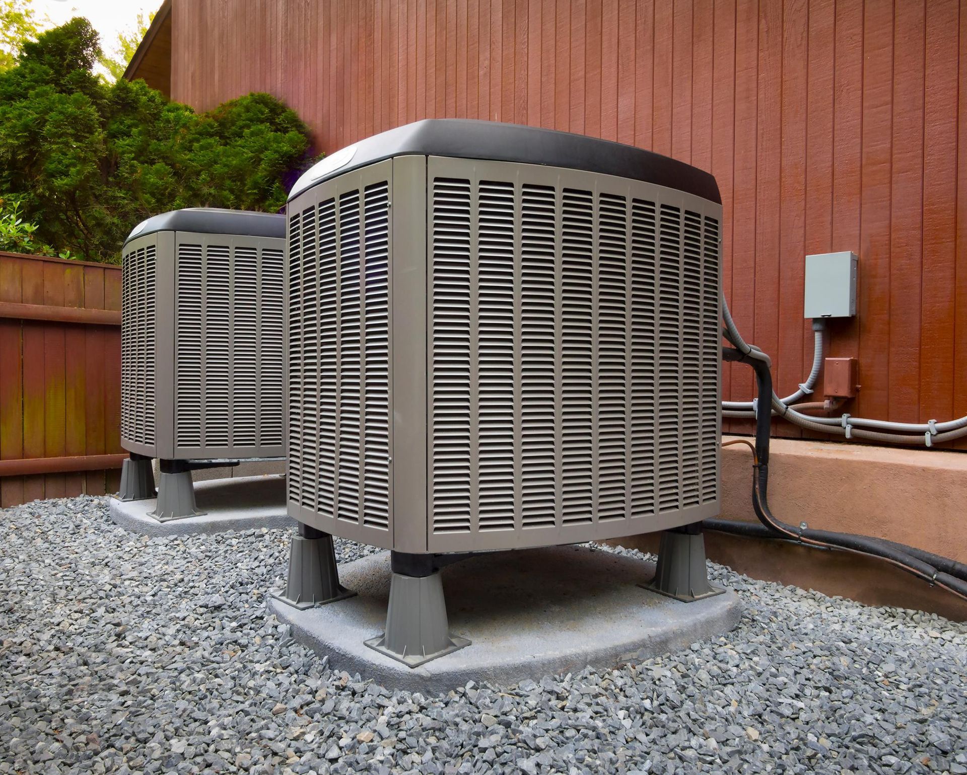 Two air conditioners are sitting on top of gravel in front of a wooden fence