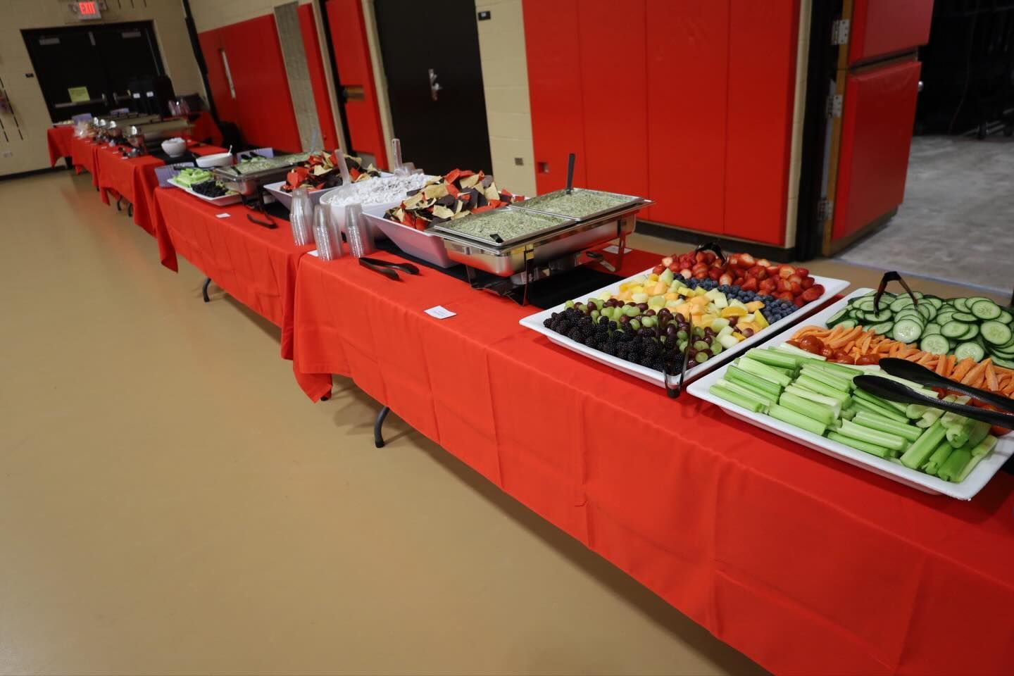 Buffet table with a variety of foods, covered with a red tablecloth, in a brightly lit room.