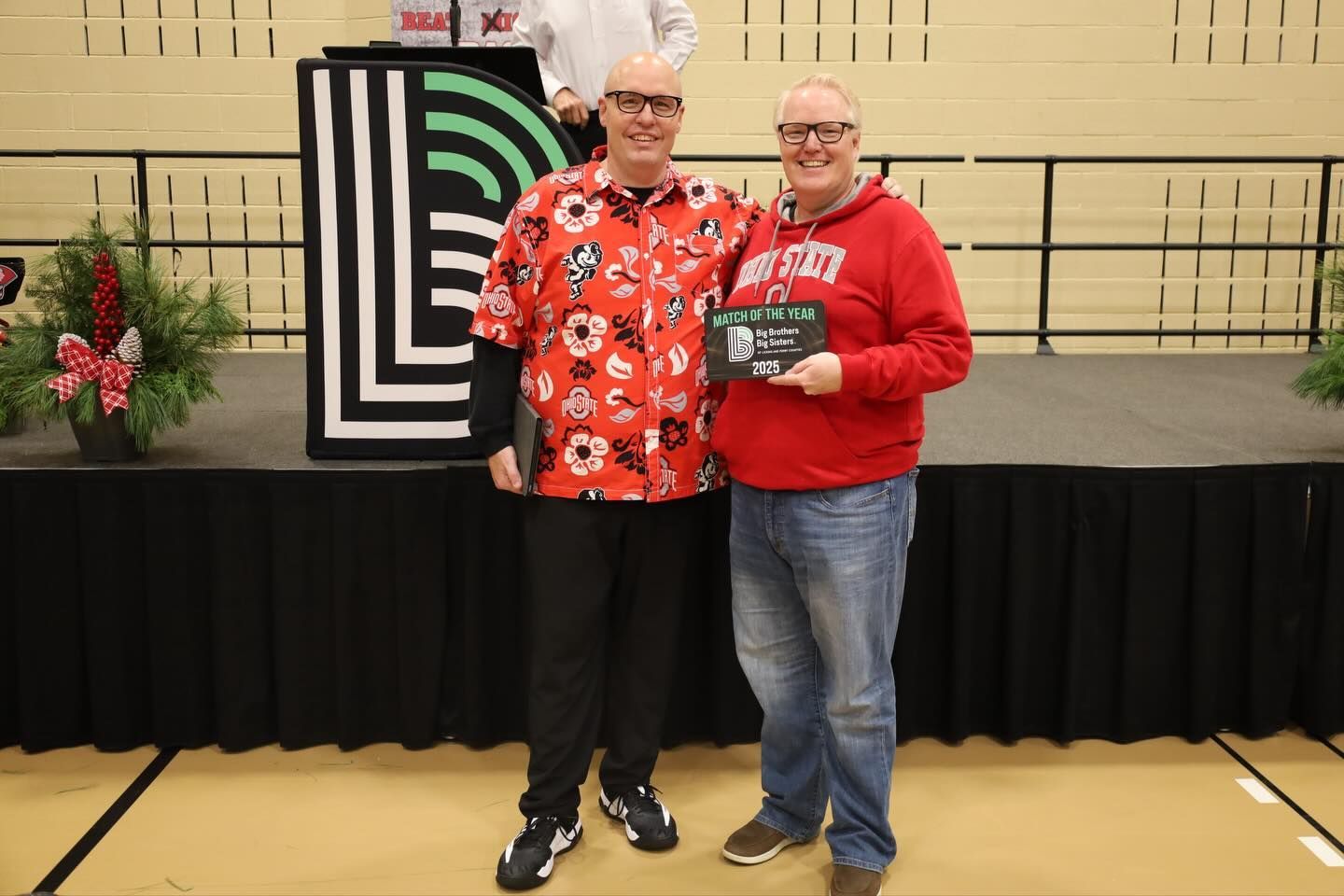 Two men stand on stage. One in a red shirt with a book, the other in a red sweatshirt.