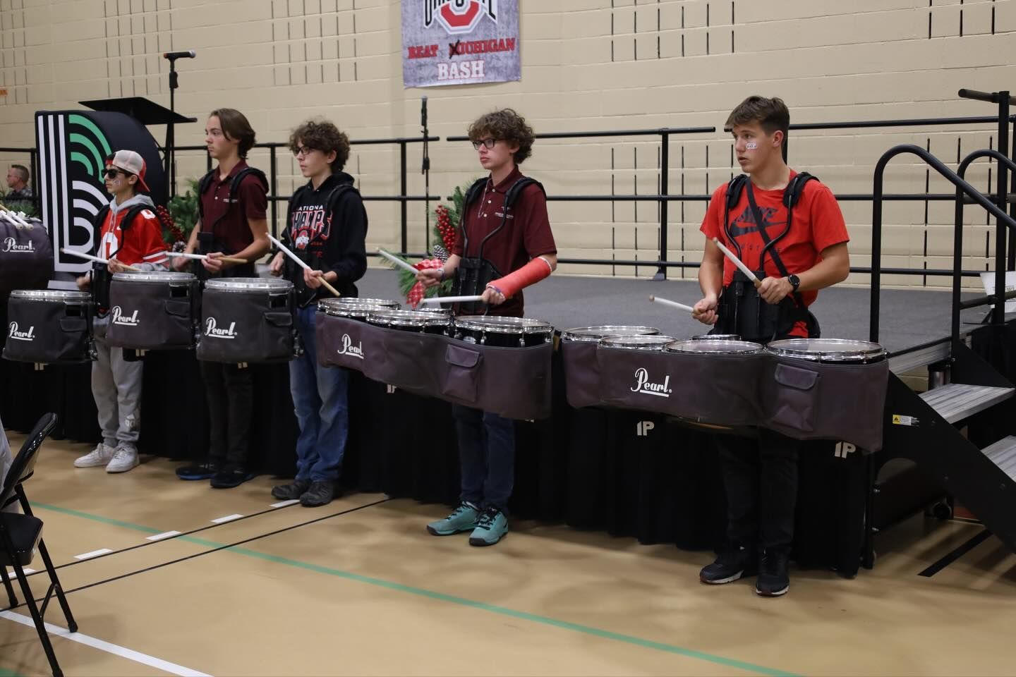 Drumline performing on stage; indoor setting. Musicians in red and black, playing drums.