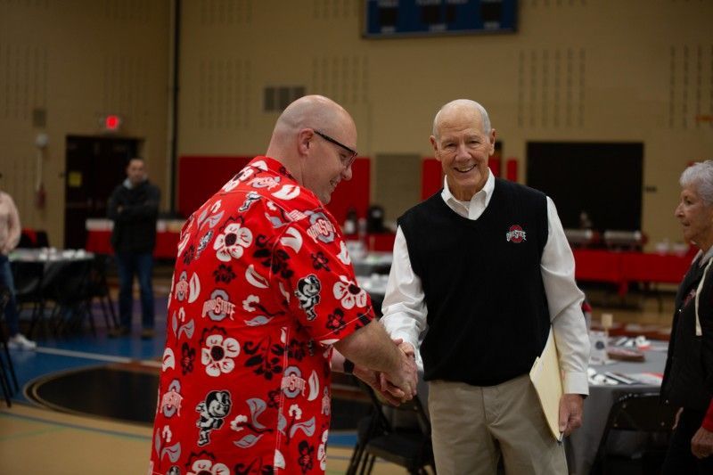 A man in a red shirt is shaking hands with another man in a black vest.