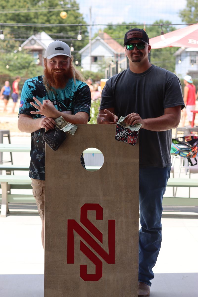 Two men are standing next to a cornhole board with the letter z on it.
