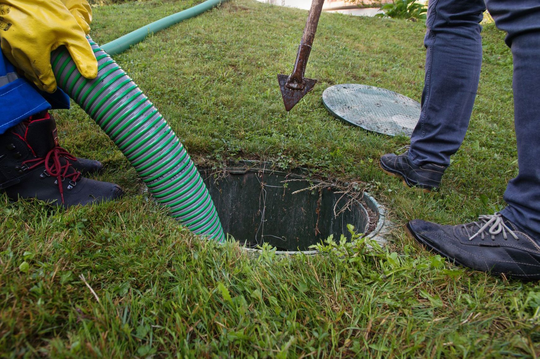 A septic tank is being serviced; a worker is holding a hose over an open tank in a grassy yard.
