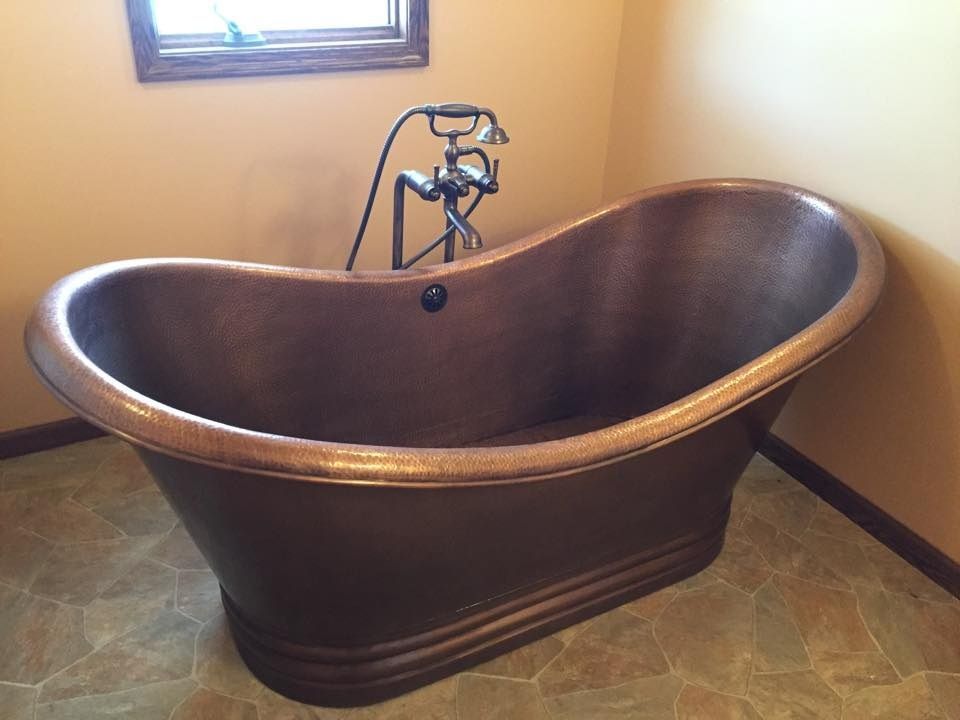 Copper bathtub in a bathroom, with a faucet. Set against tan walls and a stone floor.