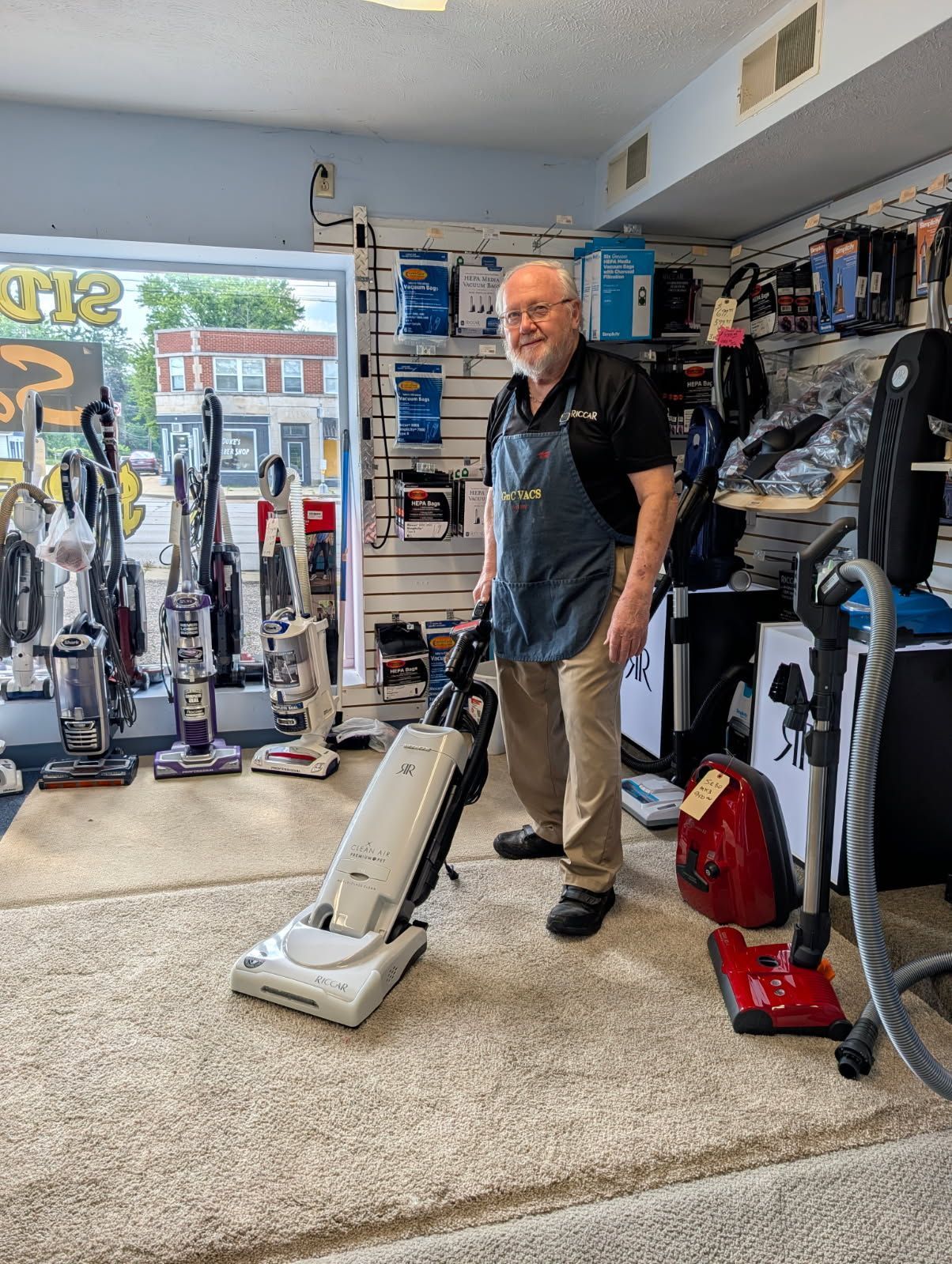 Man vacuuming carpet in a vacuum cleaner store, with upright vacuums displayed around him.