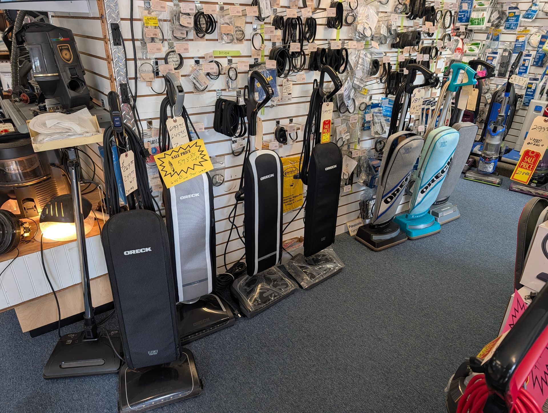 Row of upright vacuum cleaners displayed in a shop aisle, with a lit display case on the left.