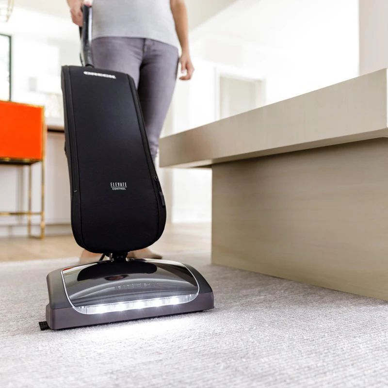 Person using a black upright vacuum cleaner on a carpeted floor indoors.