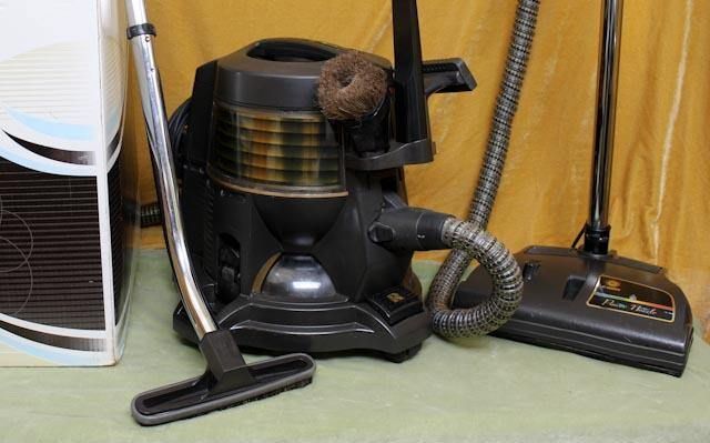 Black canister vacuum with attachments on the floor beside a white appliance and a yellow curtain.
