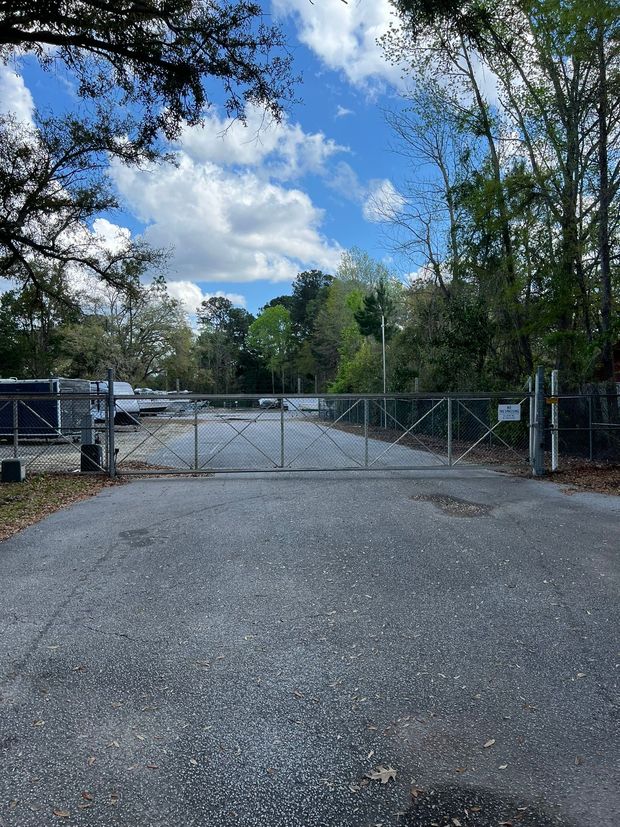 Gated asphalt road leads to open area, trees, blue sky. Metal gate closed.