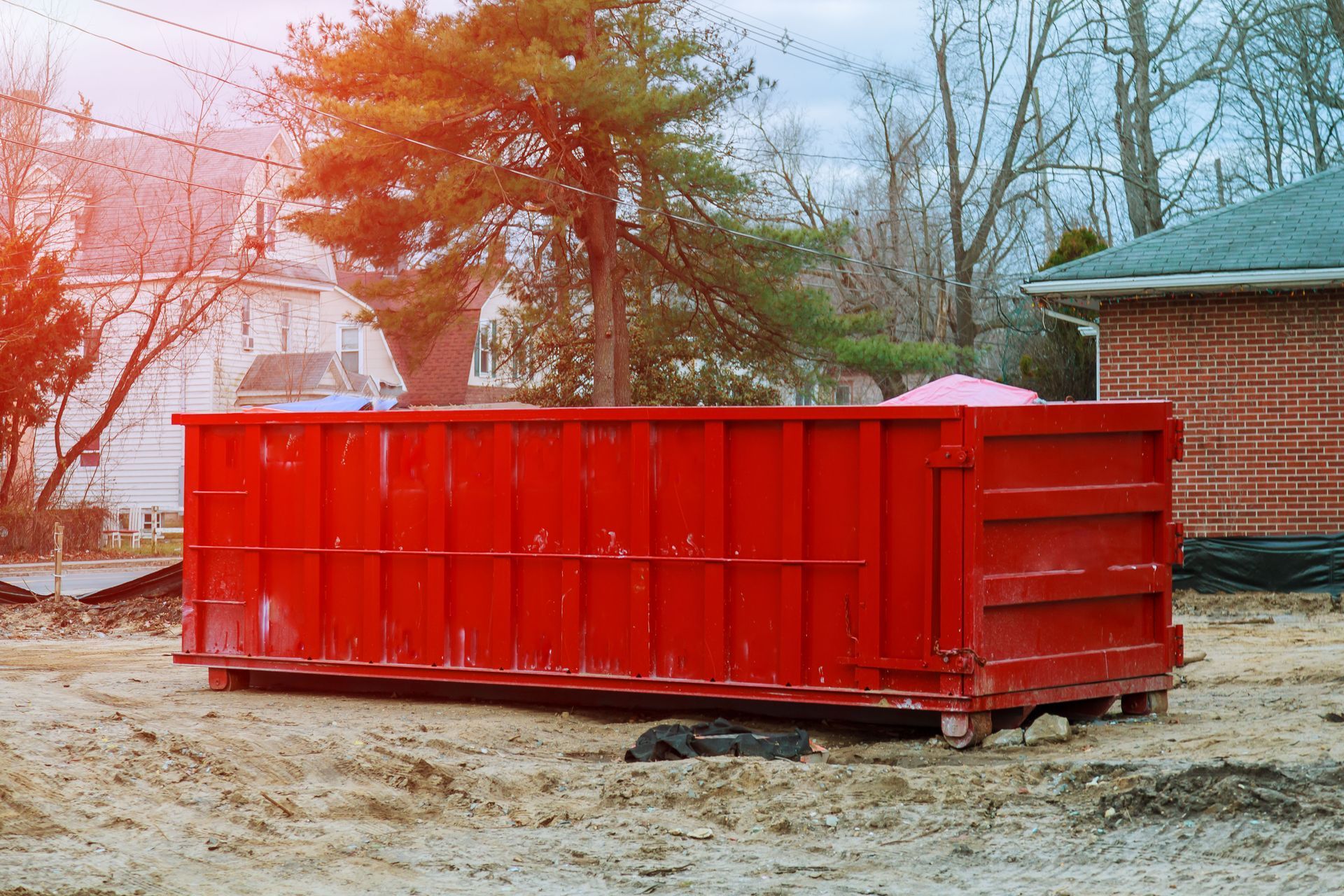 Red dumpster on a dirt lot, likely construction site, houses in the background.