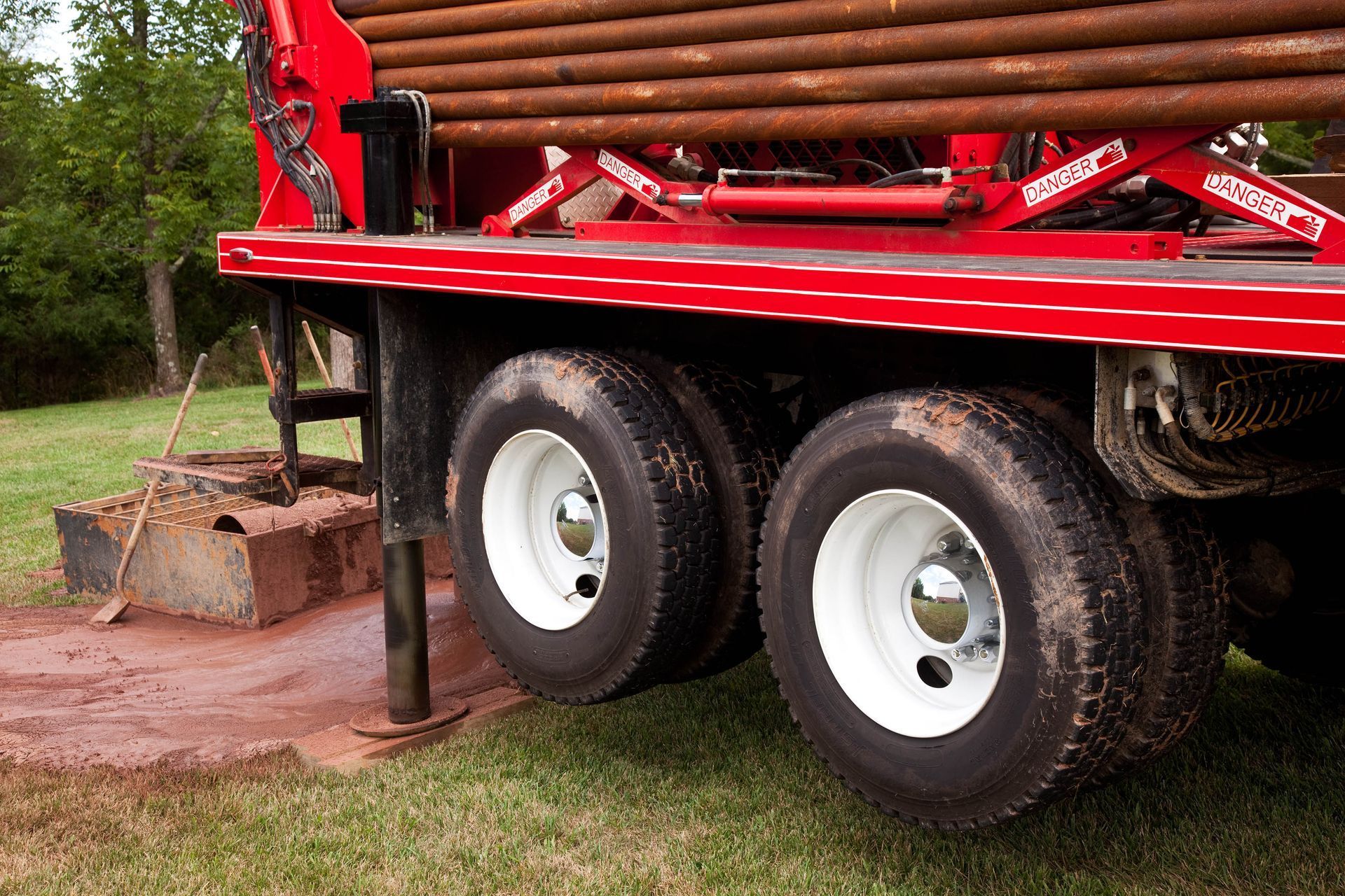 Red drilling rig with dual rear tires on grass, near a hole.