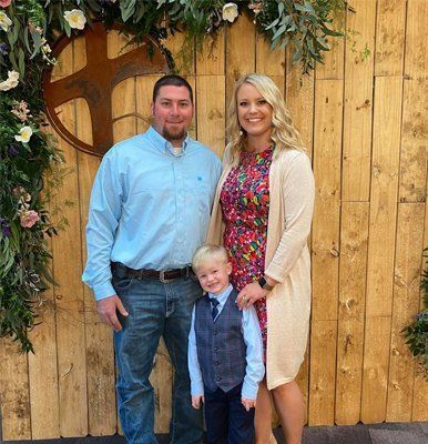 Family of three posing in front of a wooden wall with floral decor; man in blue shirt, woman in floral dress, child in vest.