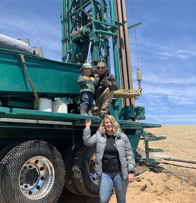Woman standing near a drilling rig, smiling. Man and child on the rig. Blue sky background.