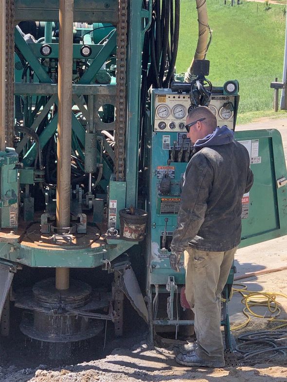 Man operating drilling equipment outdoors. Green machinery and a control panel are visible.