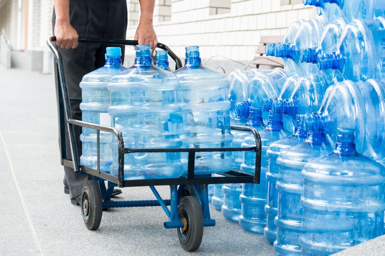Person pulling a hand cart loaded with large blue water bottles near a stack of more bottles.