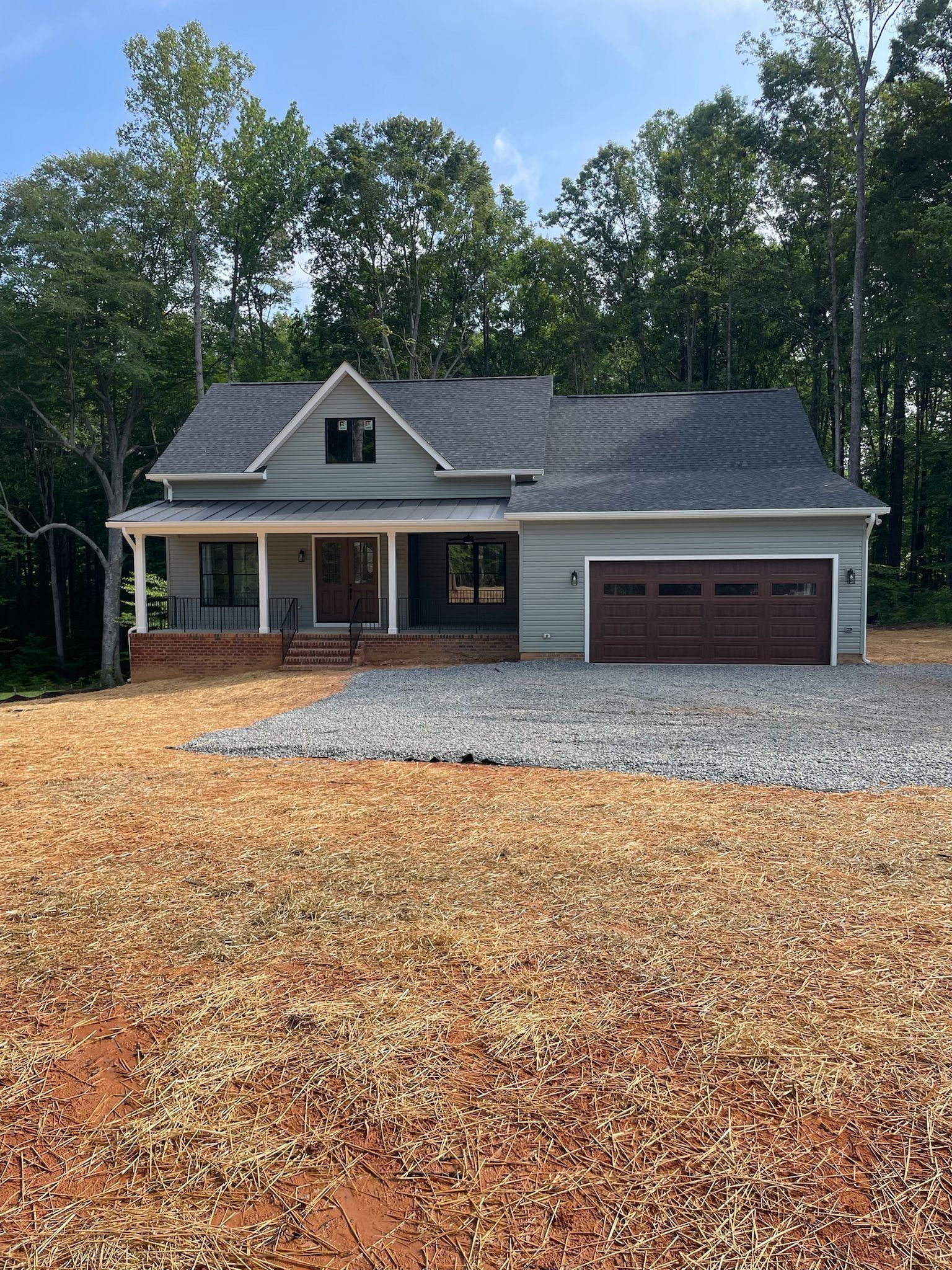 Light blue house with brown garage door, brick porch, and gravel driveway, set in wooded area.