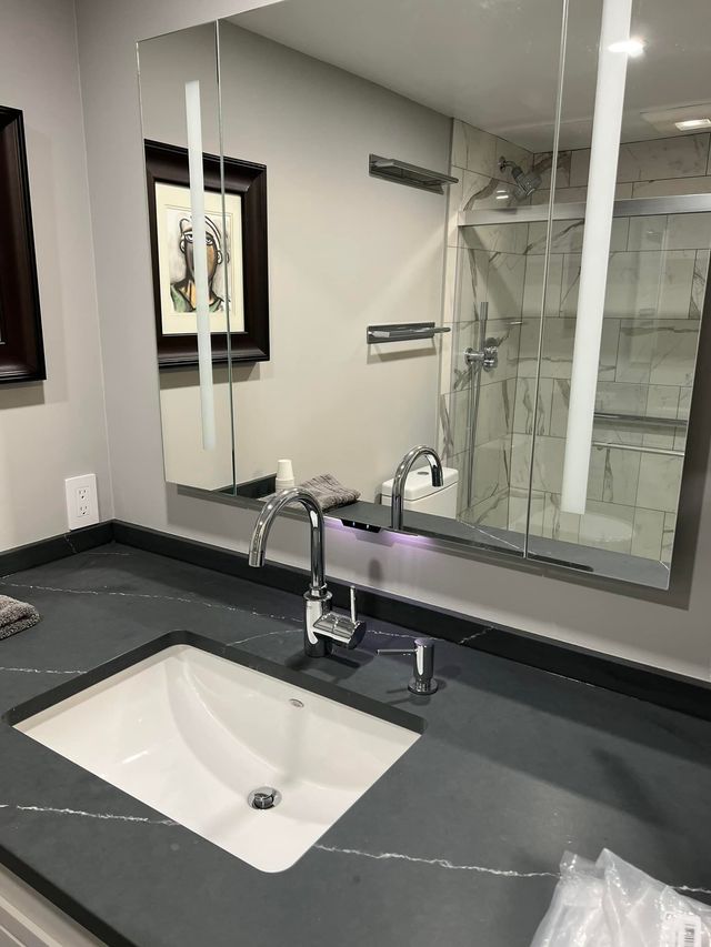 Modern bathroom sink with chrome faucet, gray countertop, and large mirror reflecting the shower area.