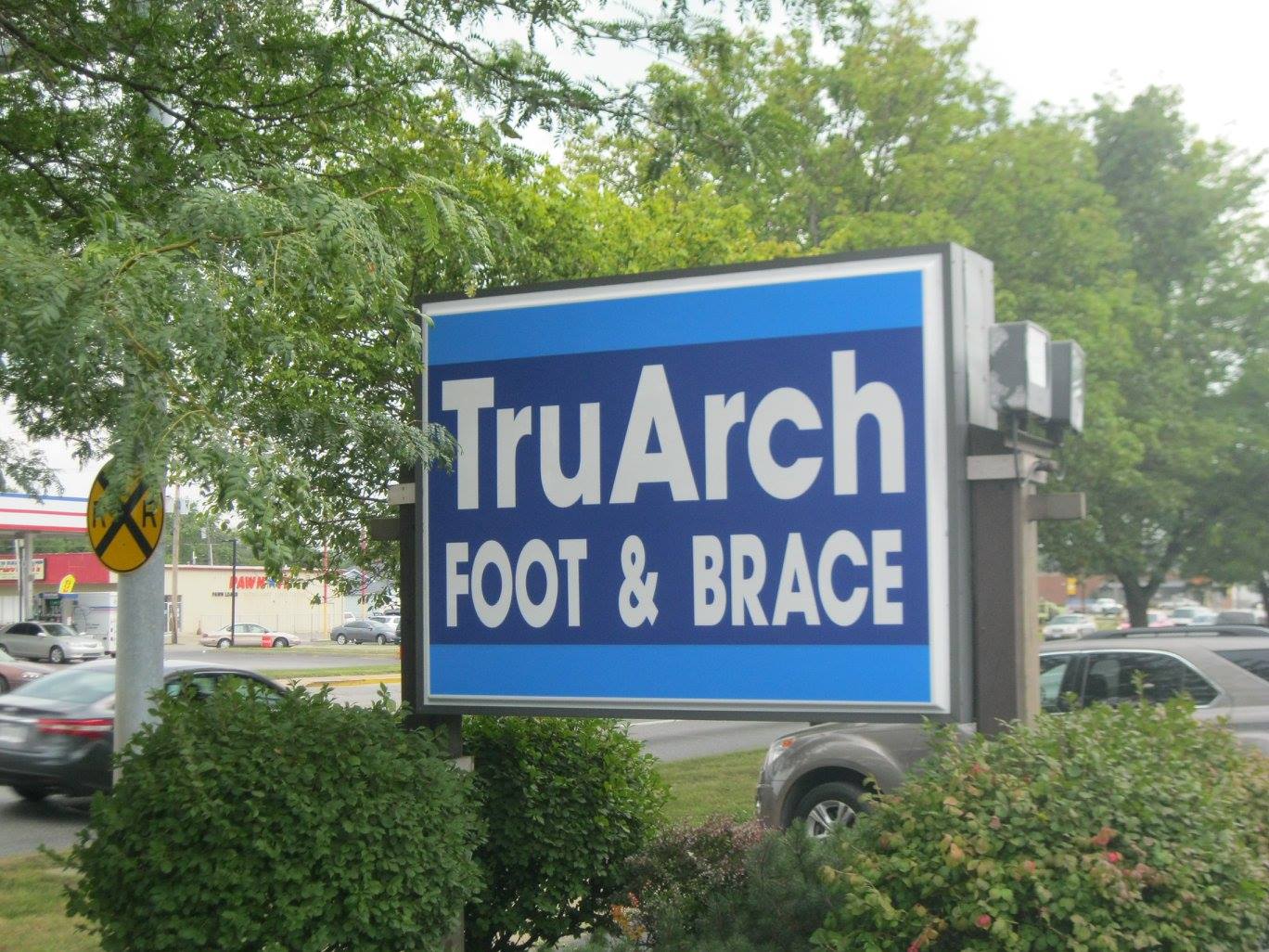 Sign for TruArch Foot & Brace, white text on blue background. Located outdoors with greenery and passing cars.