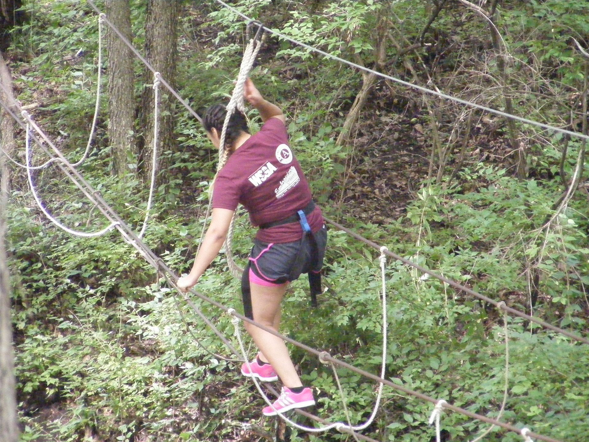 Person on a rope course, walking across a suspended bridge in a forest setting.