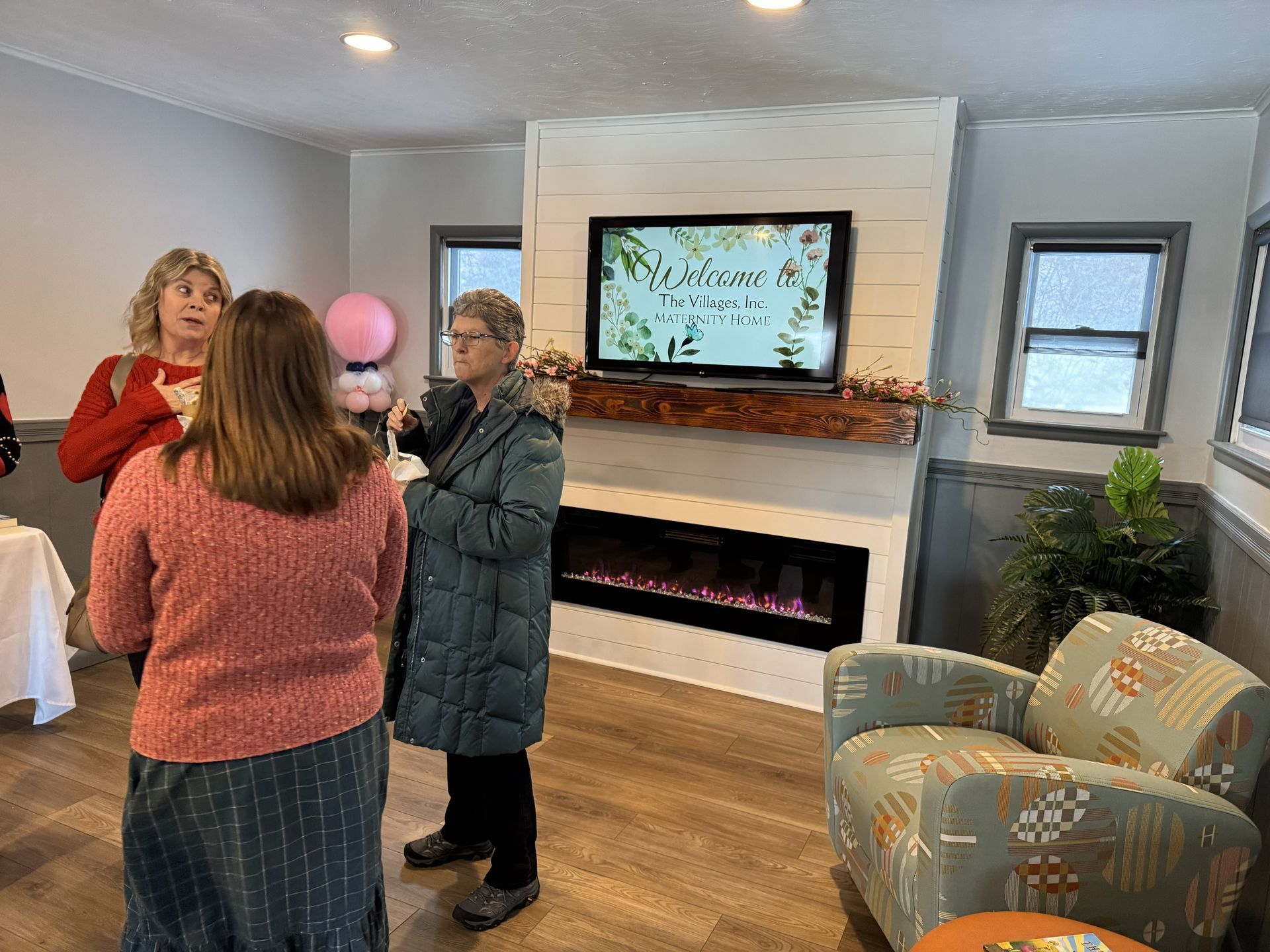 Three women talking in a well-lit room with a fireplace, TV, and patterned armchair.