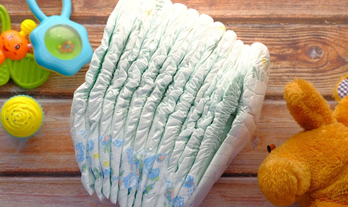 Stack of baby diapers with floral patterns, next to baby toys on a wooden surface.