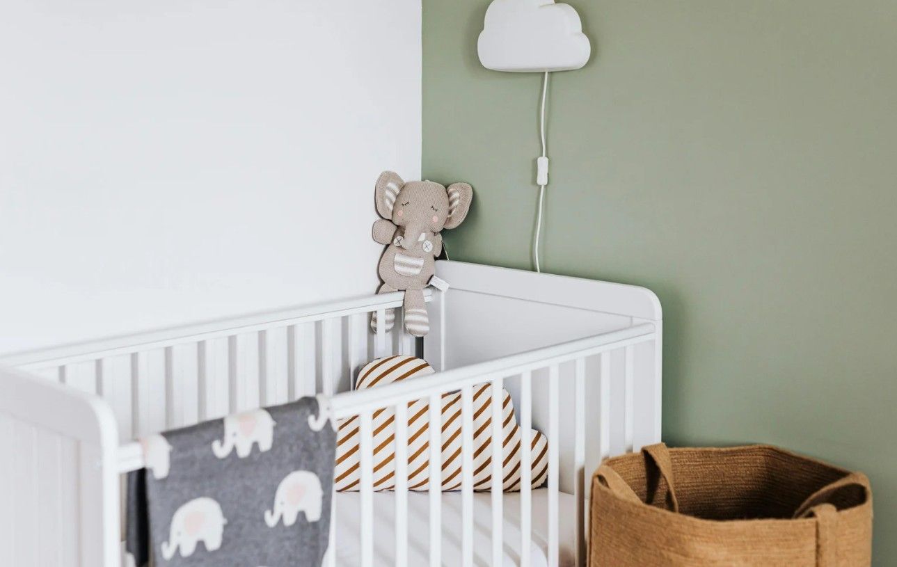 White crib in a nursery corner with a plush elephant toy, a cloud-shaped light, and a tan basket.