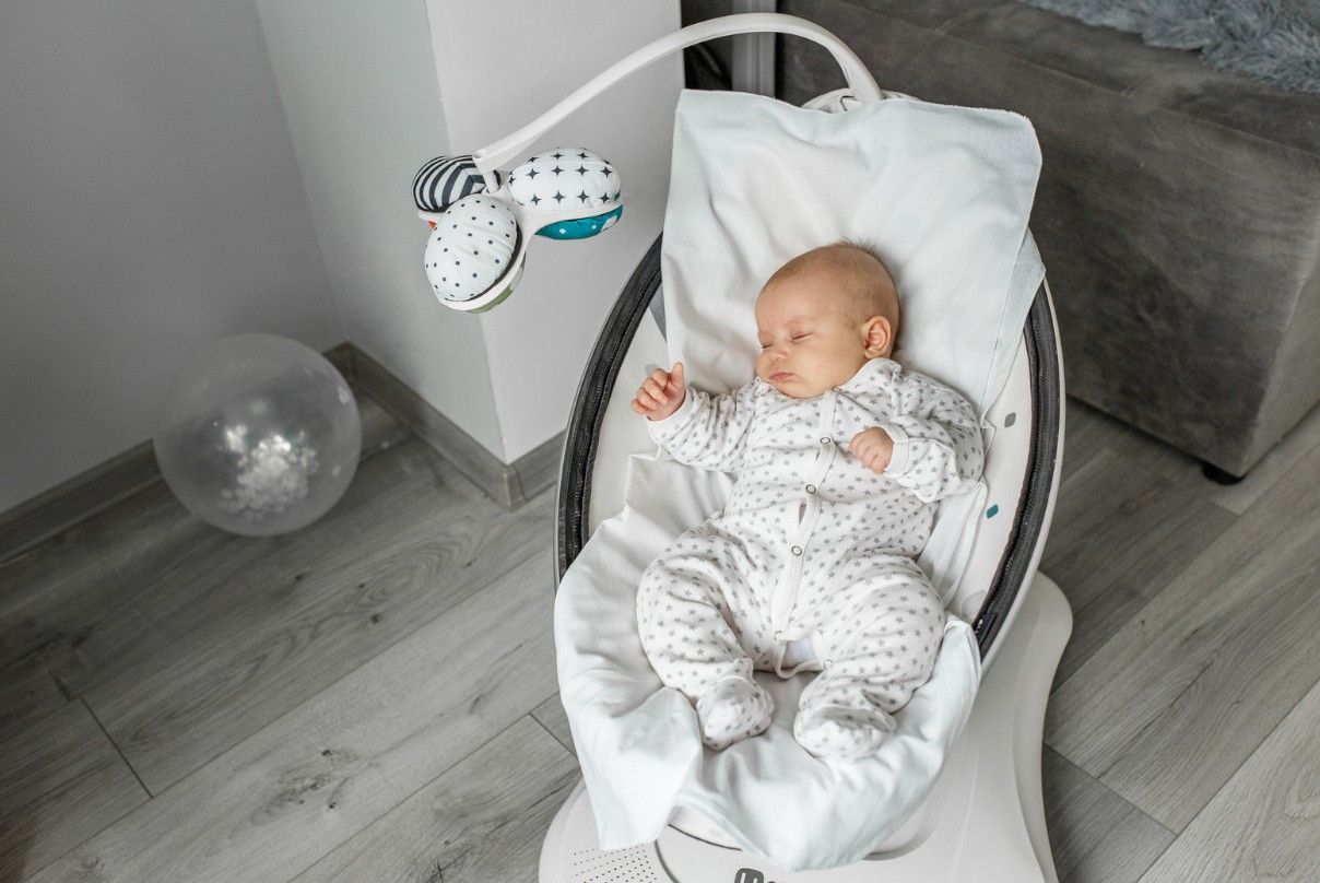 Baby sleeping in a white and gray baby bouncer, with a toy mobile overhead.