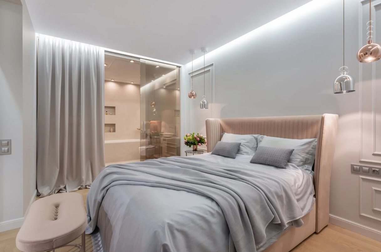 Bedroom with a bed, light-colored bedding, and a soft pink headboard. A window overlooks a bathroom.