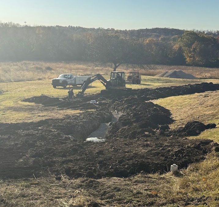 Excavator digging a trench in a field, with a truck and pile of dirt nearby. Autumn foliage in the background.