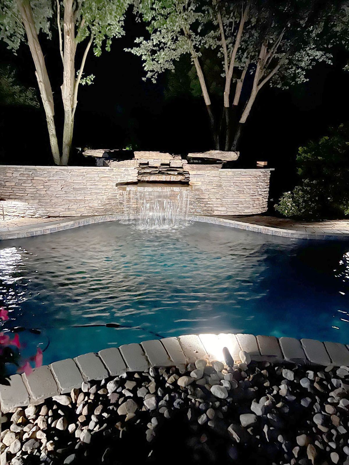 A well-lighted swimming pool with a waterfall in the backyard at night