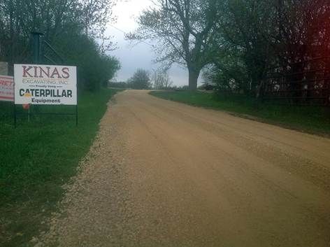 A gravel road leads into a wooded area, with a sign reading