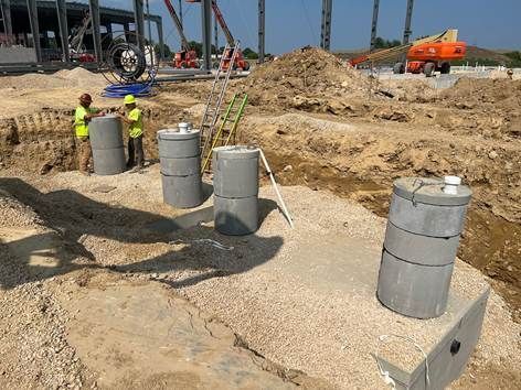Two construction workers stand by four cylindrical concrete columns at a gravel-covered industrial building site.