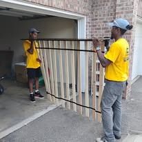 Two men in yellow shirts carrying a wooden framed object out of a garage. One man wears a cap.