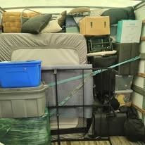 Interior of a moving truck filled with various items: furniture, boxes, and storage containers. A bed frame is prominent.