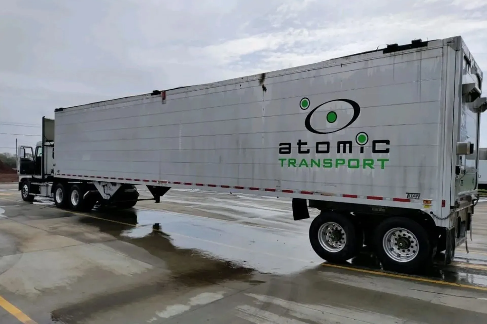 Semi-truck trailer with "Atomic Transport" logo, white with green text, parked on wet pavement.