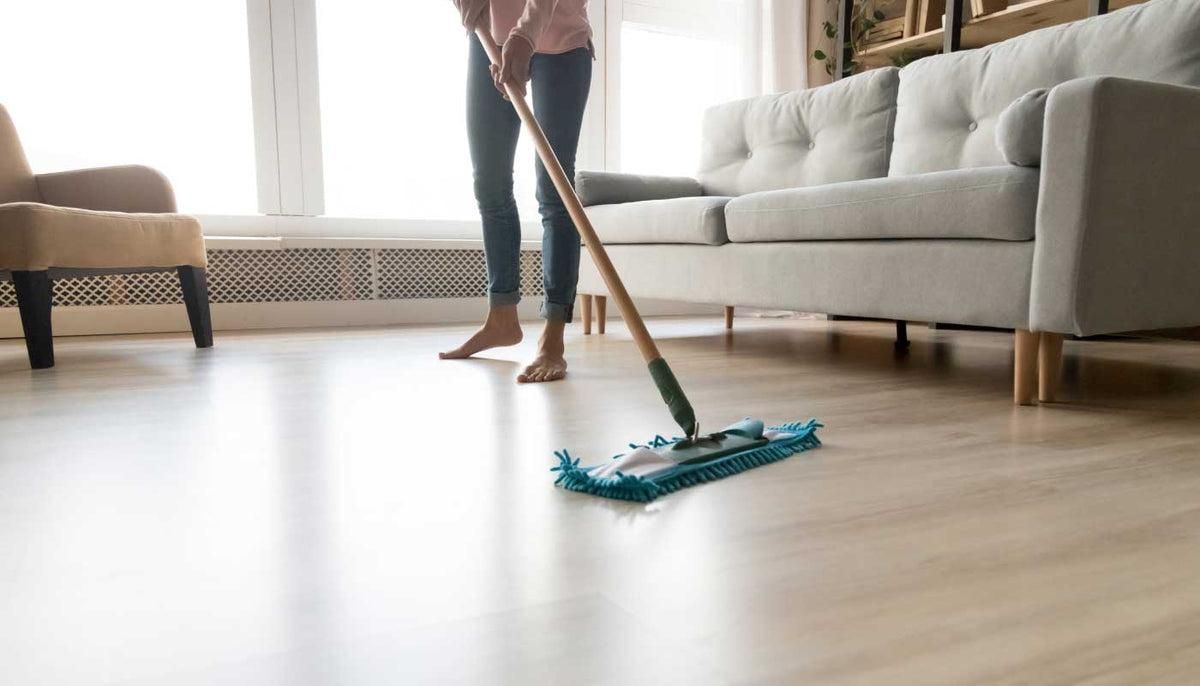 Person mopping a light-colored wooden floor in a living room, near a sofa and window.