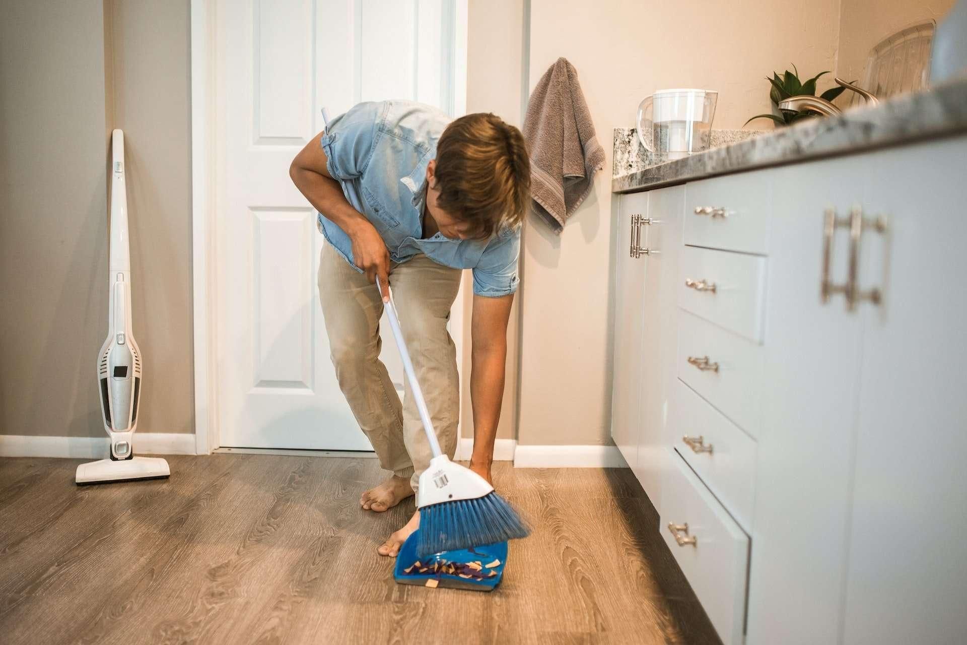 Person sweeping floor with a broom near a vacuum cleaner and white cabinets.