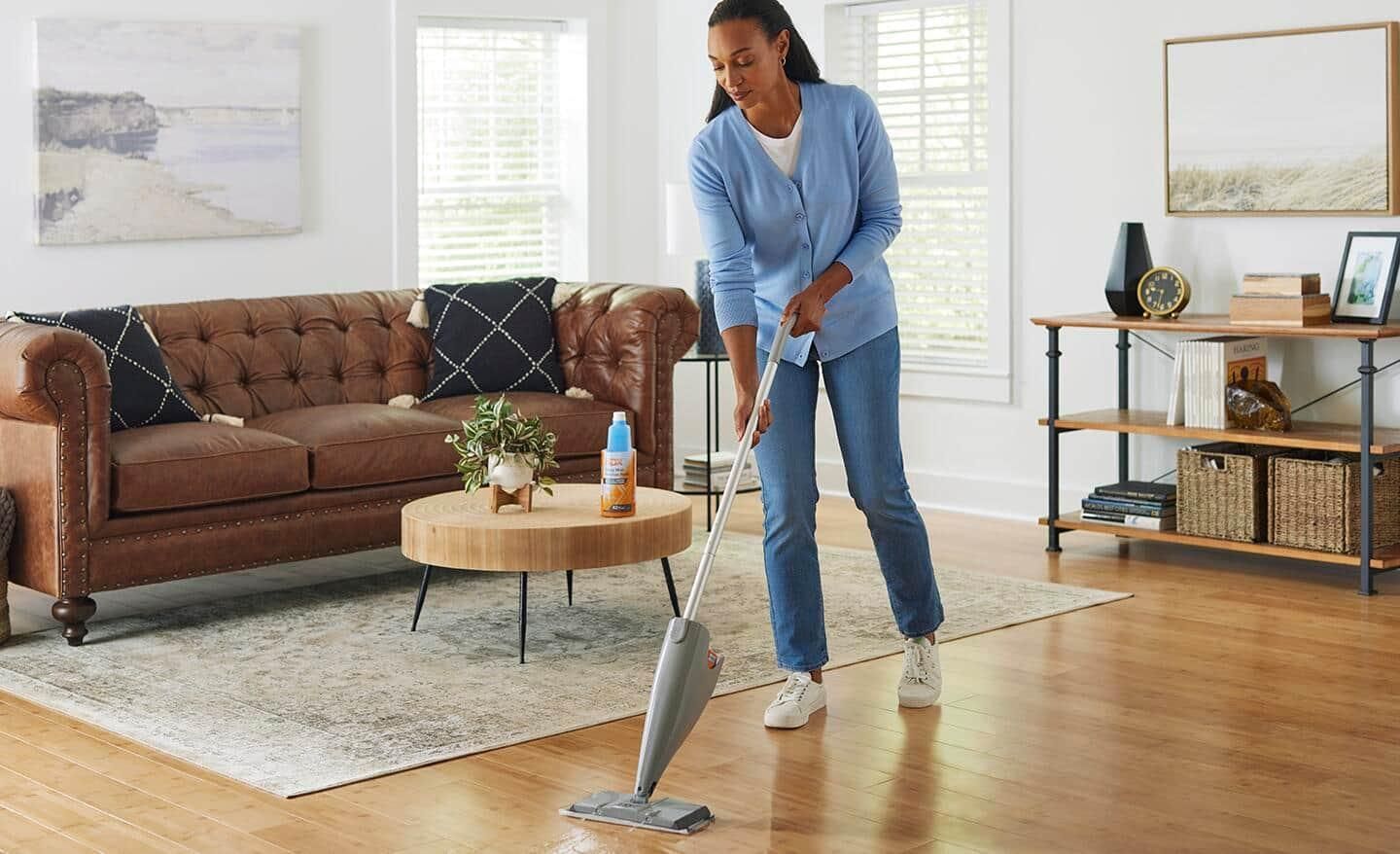 Woman mopping wooden floor in a living room, near a brown sofa and coffee table.