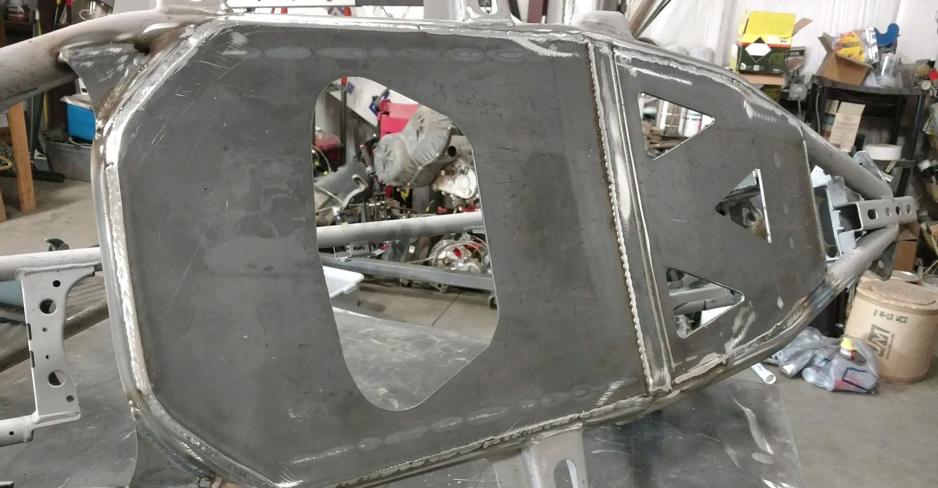 Steel frame of a vehicle body with cutouts, likely for a door and panels, in a workshop setting.