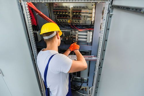 Technician in yellow hard hat wiring an open electrical control panel with tools