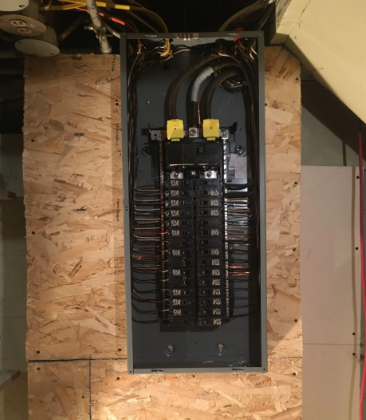 Open electrical breaker panel mounted on plywood wall with visible black cables and labeled circuits