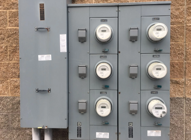 Row of electric utility meters and gray breaker boxes mounted on an exterior brick wall.