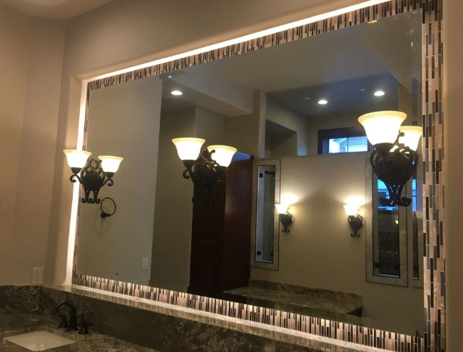 Large bathroom mirror above a sink, reflecting wall sconces and ceiling lights in a warmly lit room.