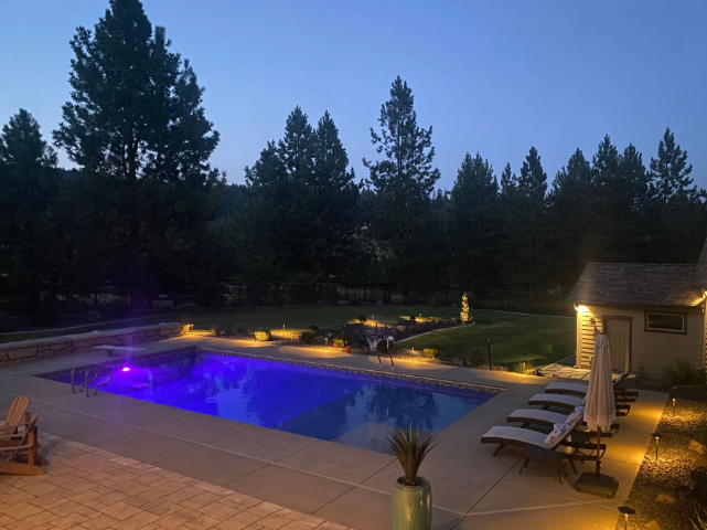 Twilight backyard with glowing purple pool, lit lounge chairs, and tall pine trees.