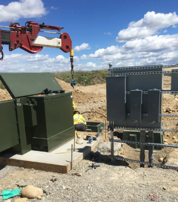 Utility equipment on a dirt lot, with a green transformer box and crane beside a black electrical cabinet under a blue sky