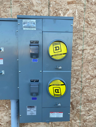 Gray electrical panel with two yellow meter dials mounted on a plywood wall